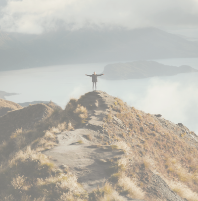 Person standing with arms outstretched on a rocky hilltop overlooking a misty lake and mountains.