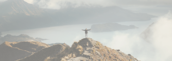 Person standing on mountain peak with arms outstretched, overlooking a foggy landscape with water and mountains in the distance.