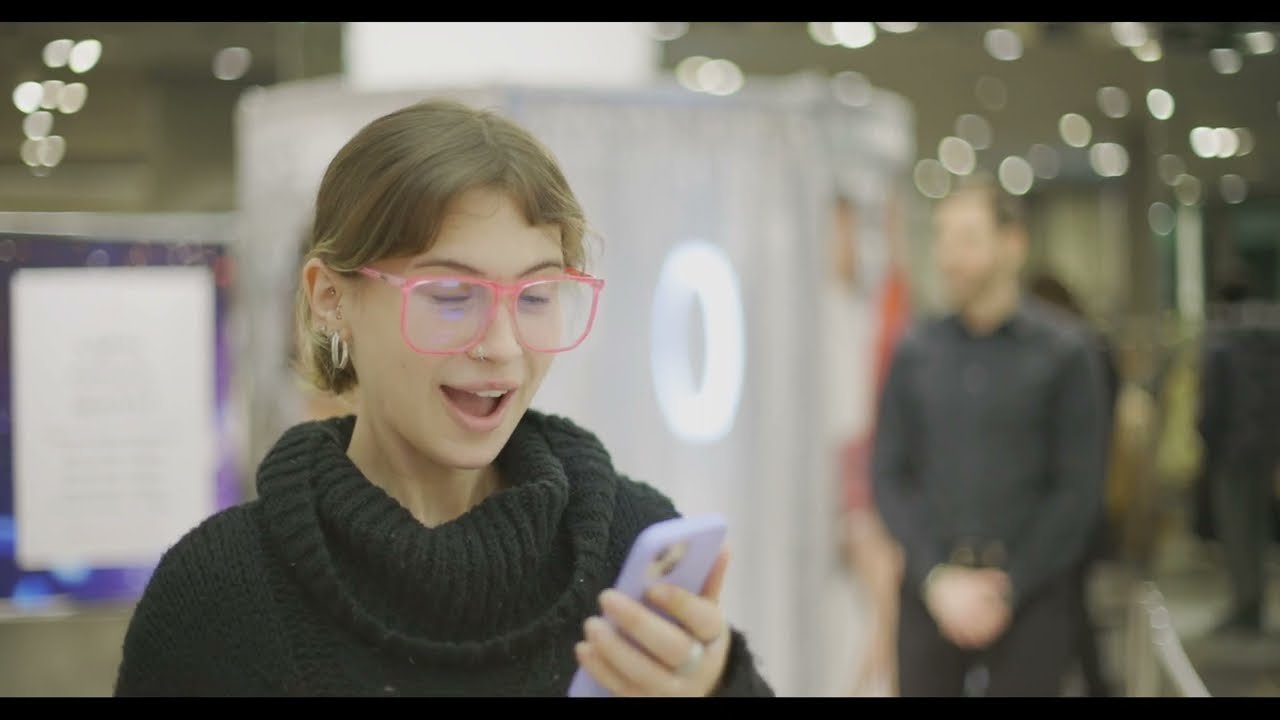 A young woman with glasses and earrings looks at her phone and smiles in a shopping mall.