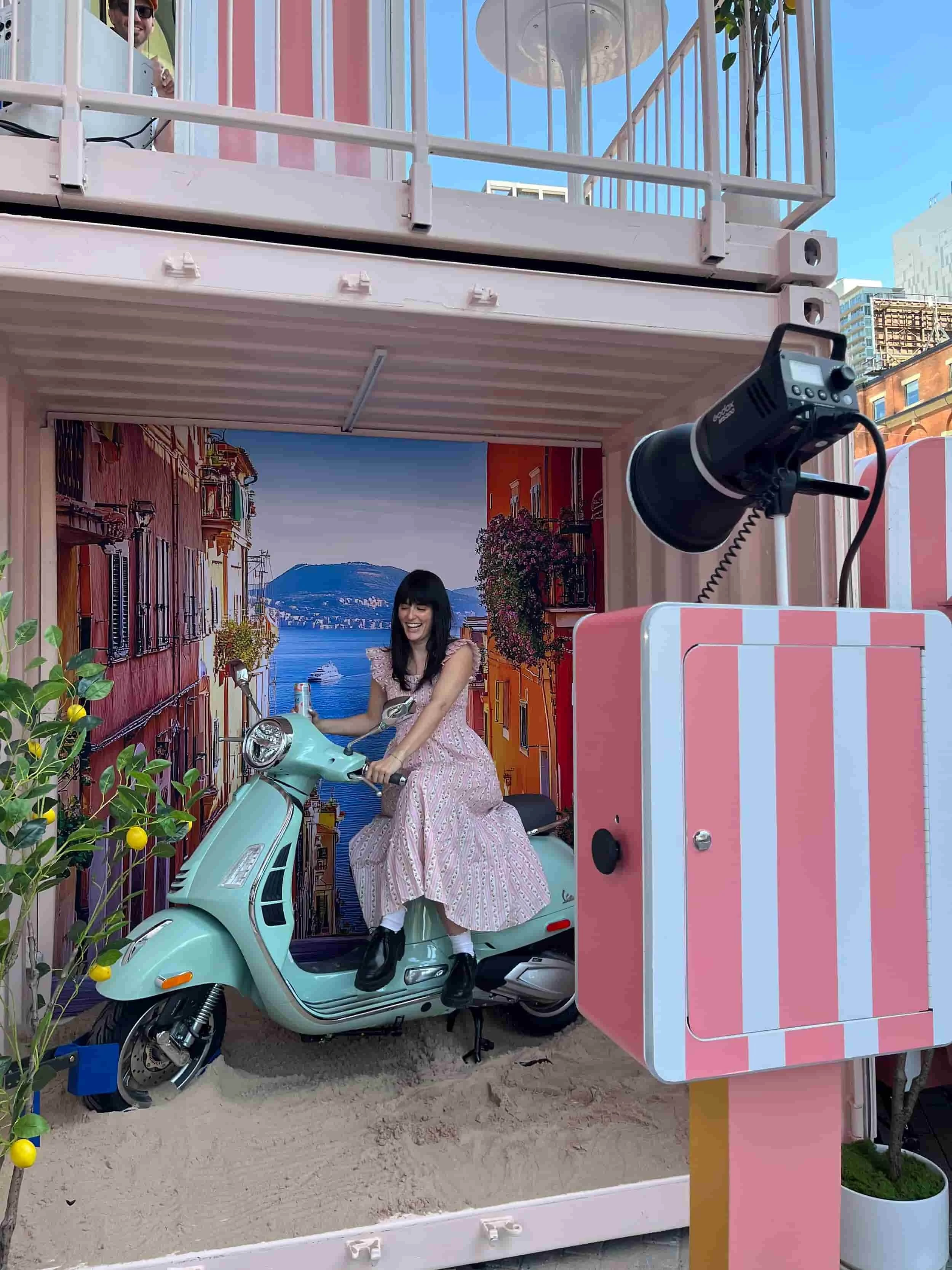 A woman wearing a pink dress is riding a mint green scooter on a miniaturized city street scene with a backdrop of a coastal city and mountains.