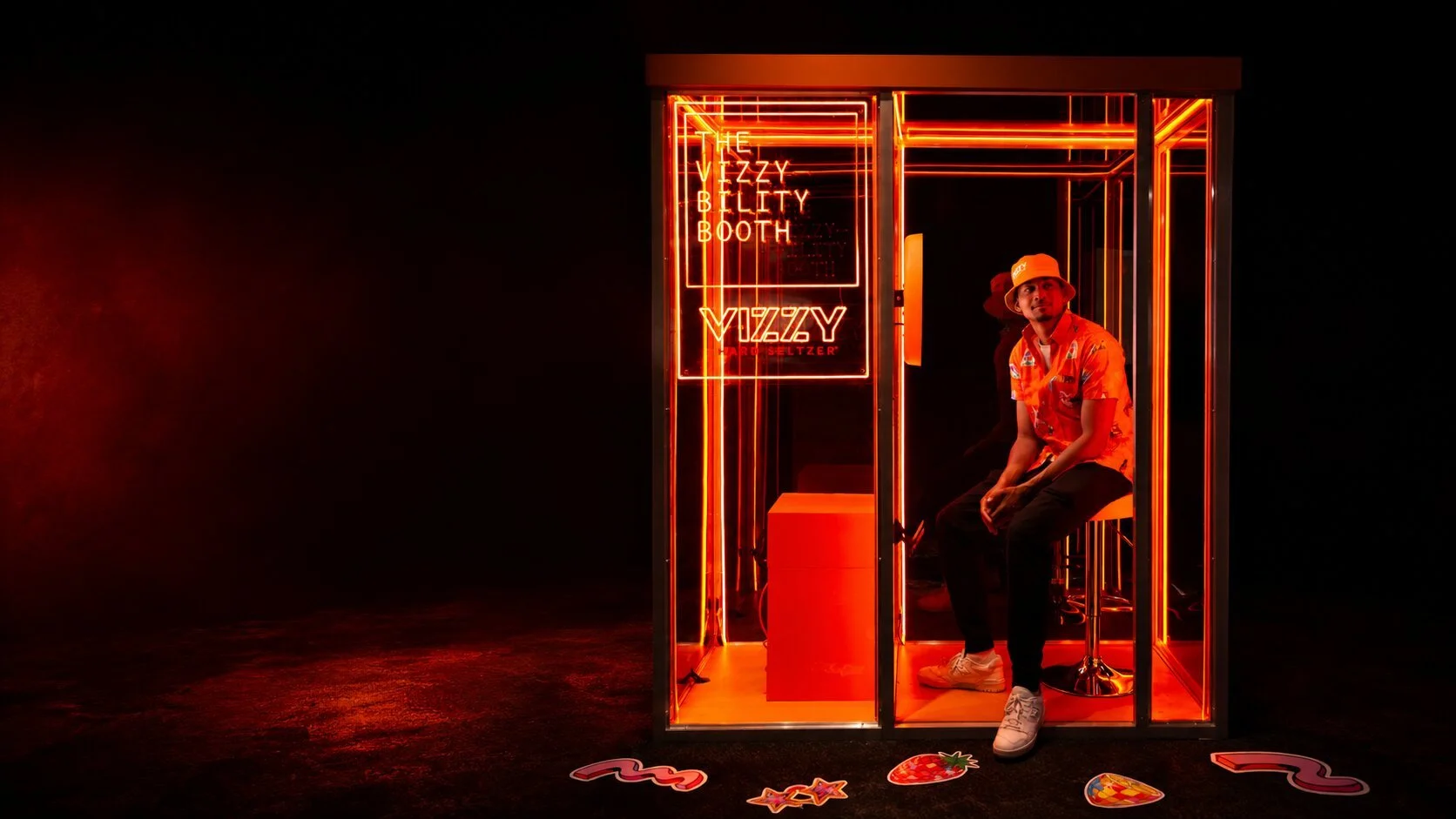 A man sitting inside a small, neon-lit booth with signs reading "The VIZZY BILITTY BOOTH" and "VIZZY" in bright red neon. The booth has a glass door and illuminated framework. The man is wearing a floral shirt, white sneakers, and a bucket hat, and is surrounded by colorful stickers on the dark floor outside the booth.