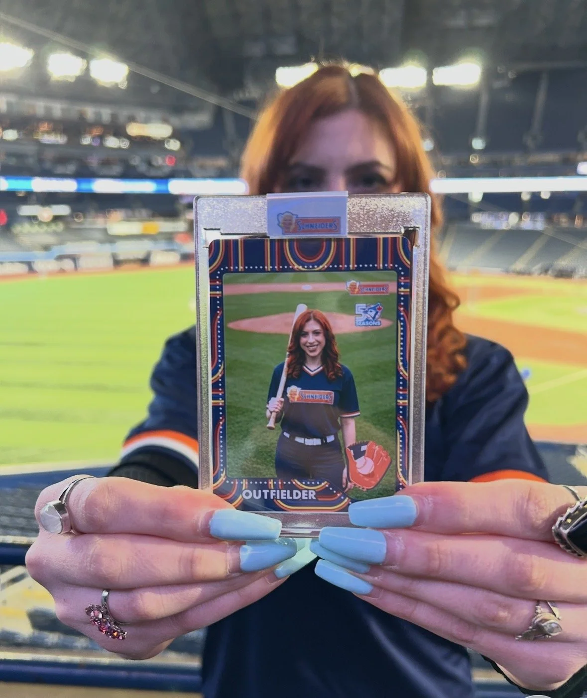 A woman with red hair holding a baseball card at a baseball stadium. The card features her smiling and dressed as a baseball player, labeled as an outfielder, with a baseball bat and glove in the background.