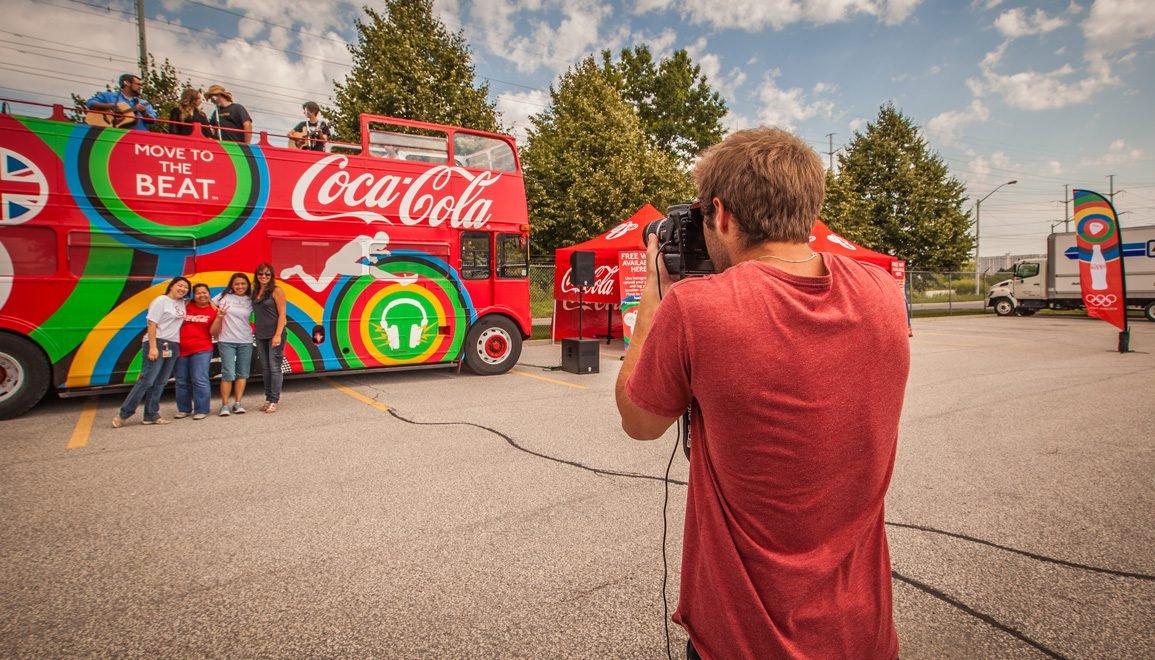 People posing for a photo in front of a colorful Coca-Cola themed bus at an outdoor event, with a photographer taking their picture.