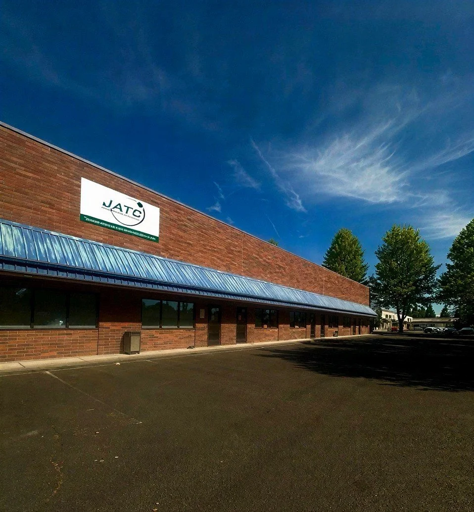 A brick building with a blue metal awning and a parking lot in front. There is a sign on the building with the logo 'JATC' and some small trees on the right side, with a blue sky and clouds overhead.