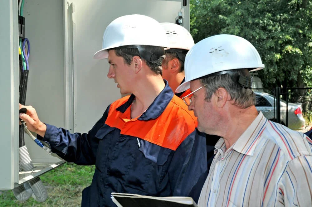 Three electricians in safety helmets working on an outdoor electrical panel, one holding a tool and the others observing, with trees and a parked car in the background.