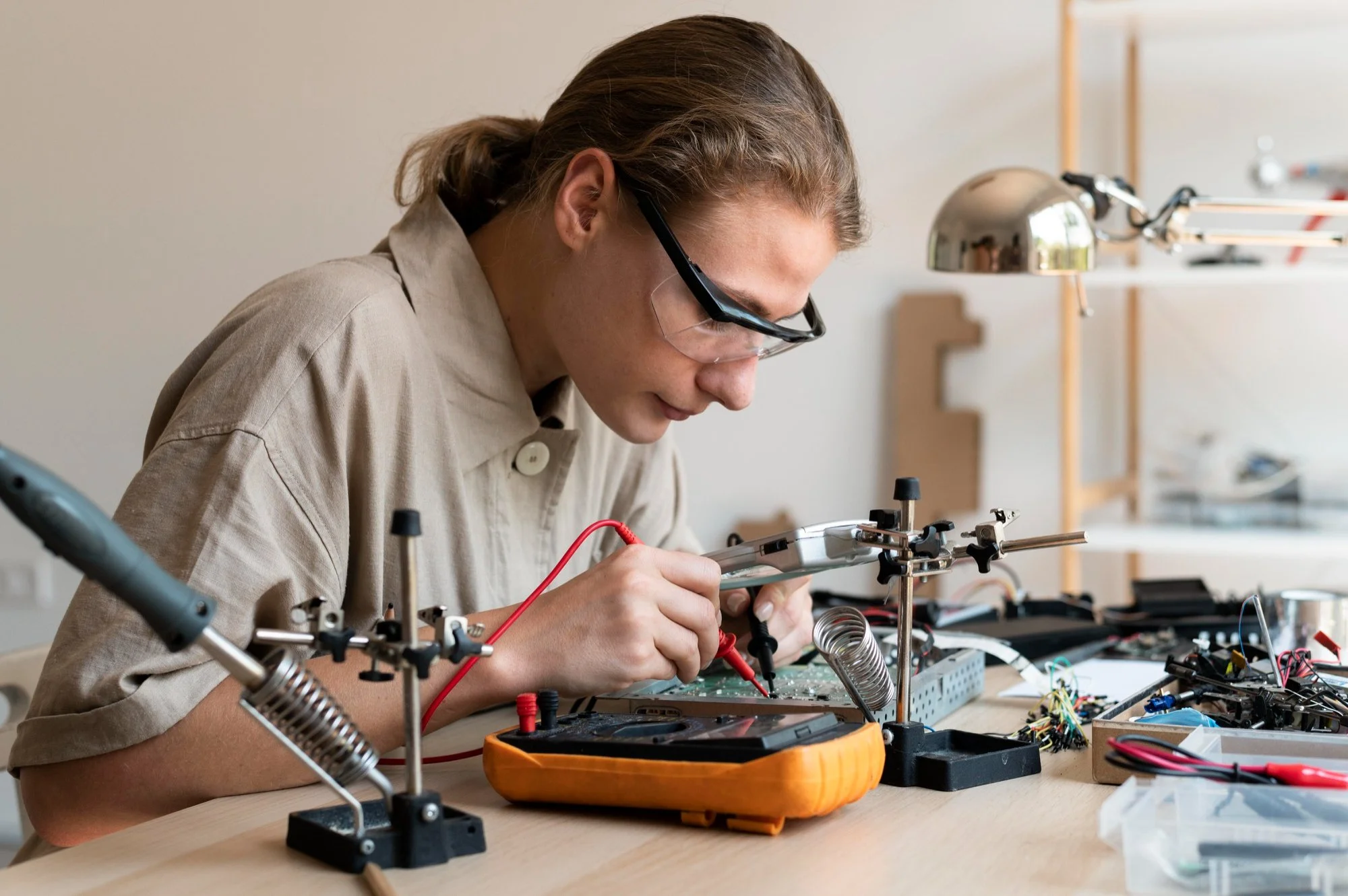 Person working on electronic circuit board at a workstation with tools and equipment, wearing safety glasses.