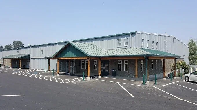 A large light gray commercial building with a green metal roof and a covered entrance with wooden beams, surrounded by a parking lot with white striped parking spaces and green posts.