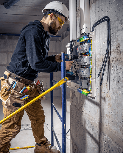 Electrician working on an electrical panel with tools, wearing safety gear and standing on a scaffolding.
