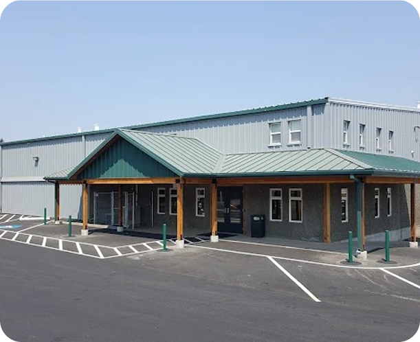A modern commercial building with a green metal roof and a covered entrance supported by wooden beams. There is parking in front with marked spaces and green posts.