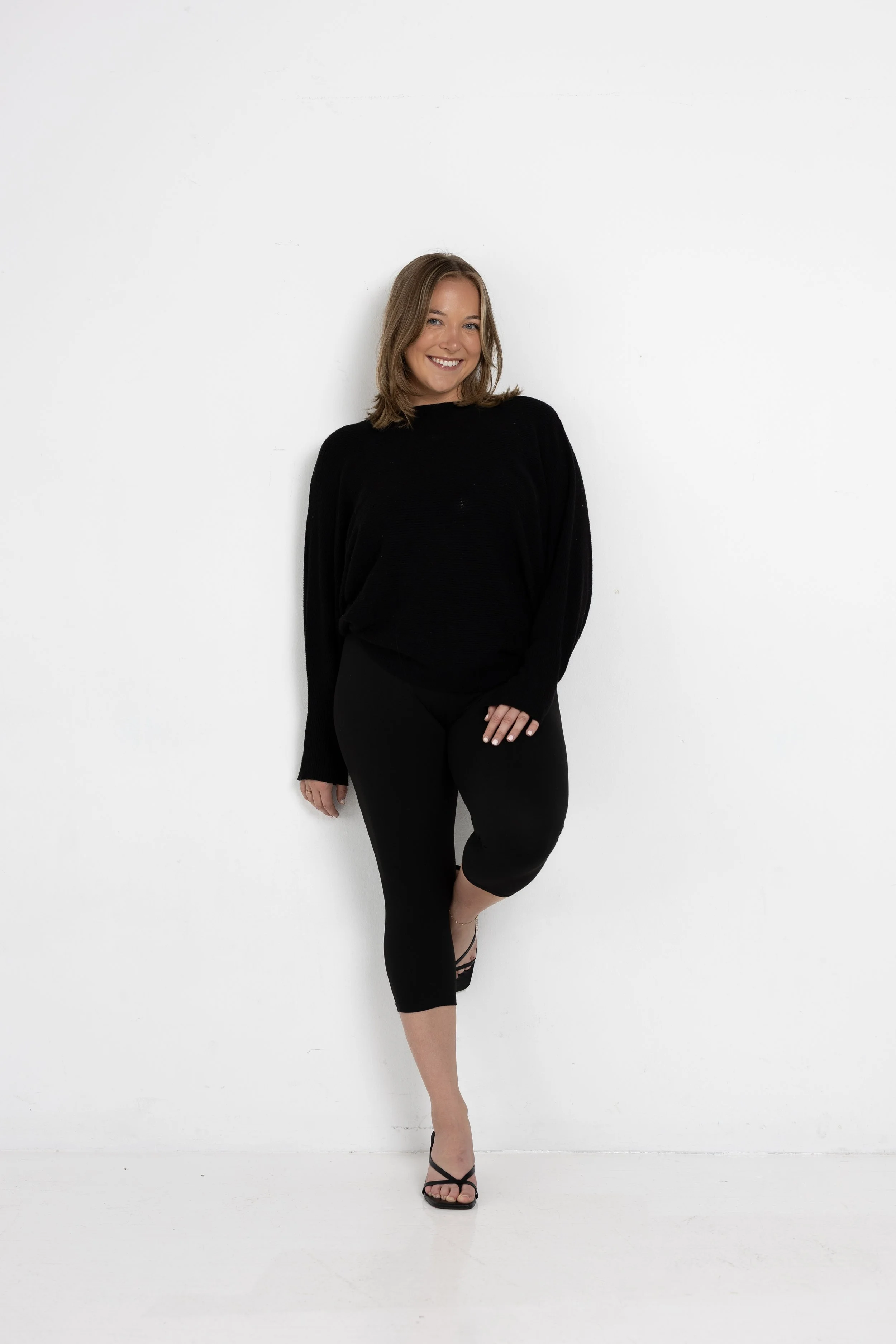 A young woman with shoulder-length brown hair smiling while standing against a white wall. She is wearing a black long-sleeve top, black leggings, and high-heeled sandals, with one leg slightly raised and her hands relaxed at her side.