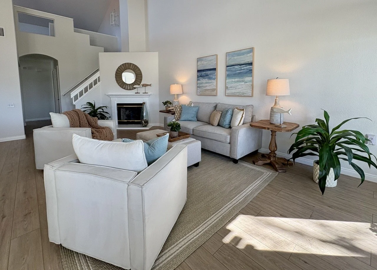 Living room with white sofas, a wooden coffee table, paintings of ocean scenes, lamps, green plants, and a fireplace.