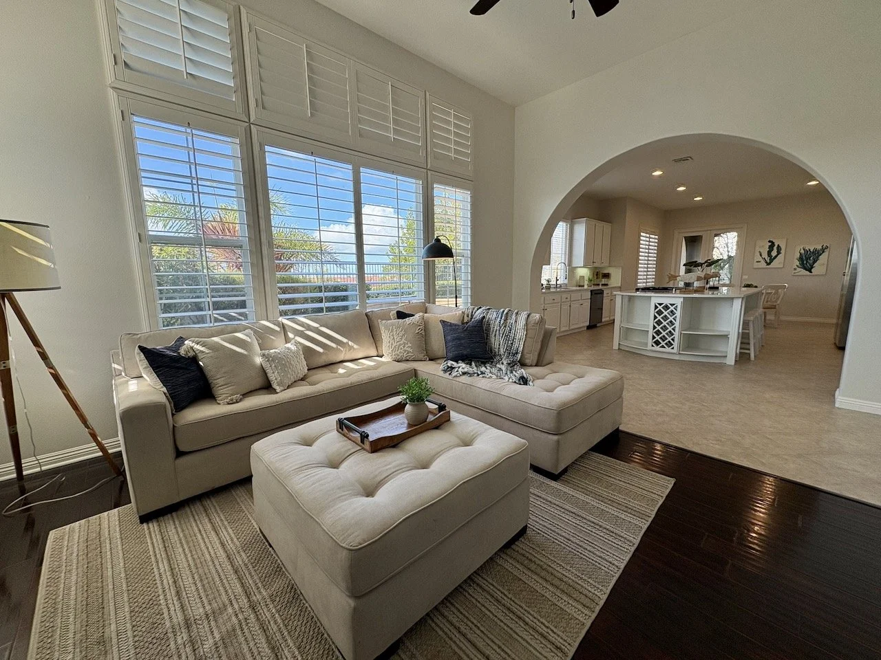 Living room with large windows, beige sectional sofa, ottoman with tray and plant, on a striped rug, open kitchen and dining area in the background with white cabinets and a curved archway.
