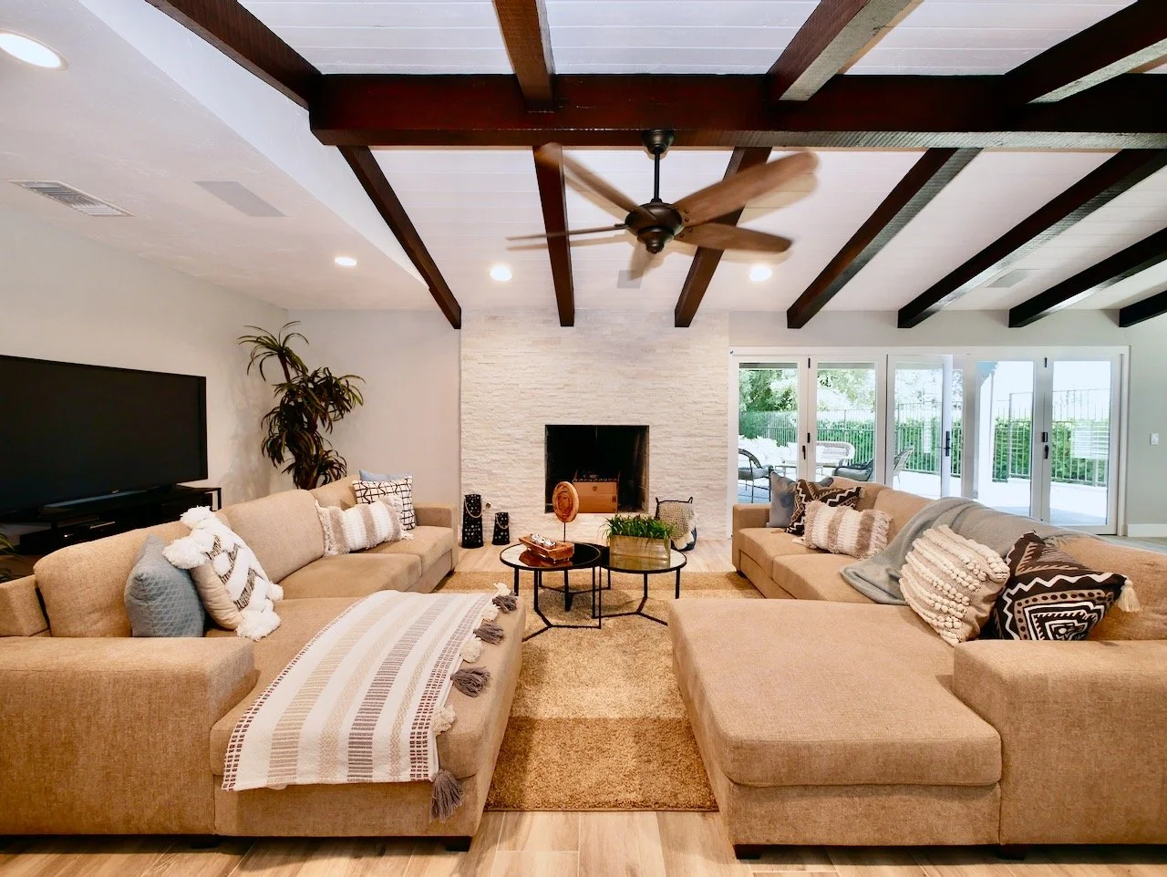 Living room with two beige sofas, a stone fireplace, and sliding glass doors leading outside. A ceiling fan hangs from the white ceiling with dark wooden beams, and there are decorative pillows and blankets on the sofas. A potted plant sits next to a TV.