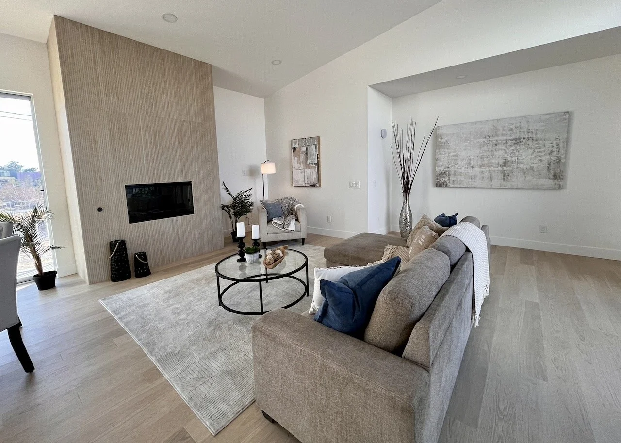 Living room with a beige sofa, artwork on a white wall, a modern fireplace, a round glass coffee table, decorative pillows, a large vase with tall sticks, and a beige armchair.