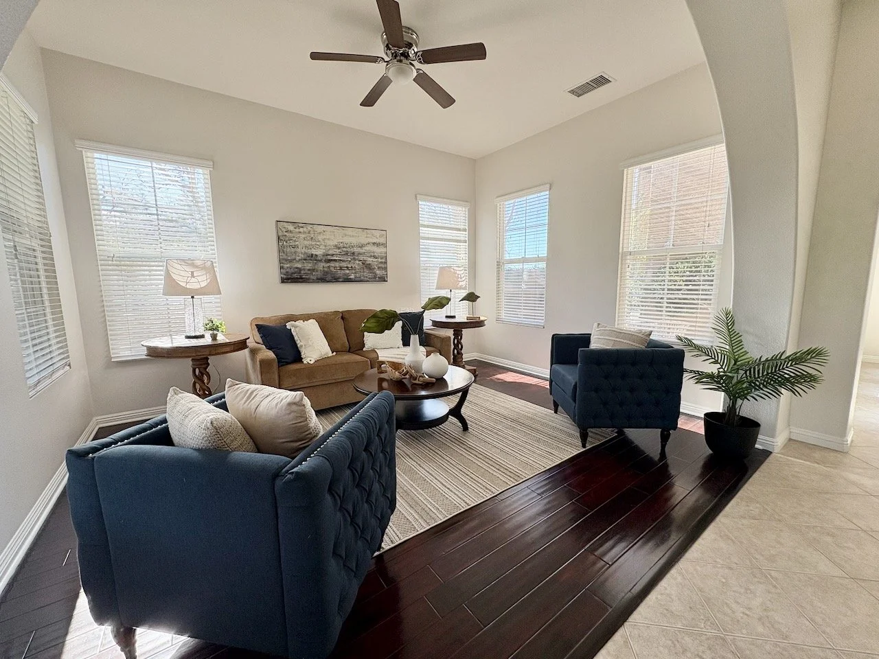 Living room with a beige sofa, two blue armchairs, a round wooden coffee table, two side tables with lamps, window blinds, a striped rug, a ceiling fan, a potted plant, and wall art.