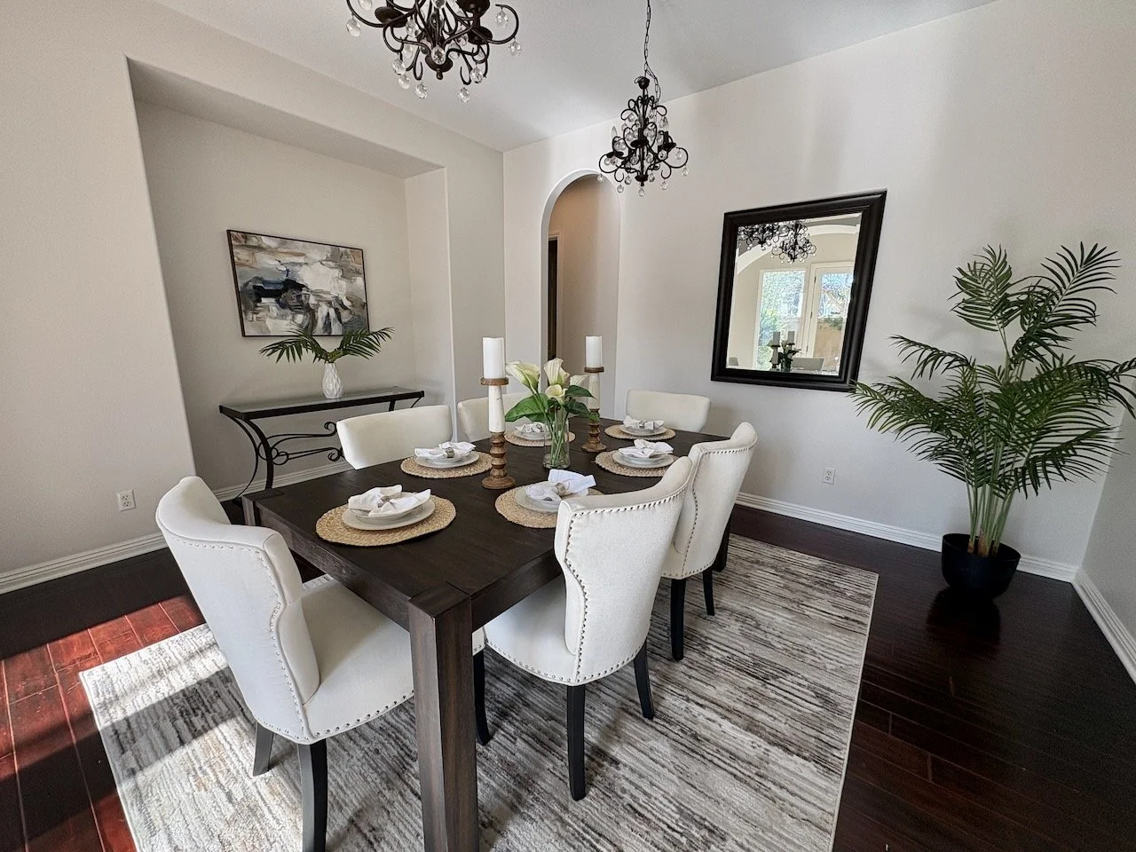 A dining room with a dark wooden table set with six place settings, each with a white napkin, plate, and woven placemat. White upholstered chairs surround the table. Two black chandeliers hang from the ceiling. There is a large mirror on the wall, a framed abstract painting, a side table with a potted plant and a vase, and a large potted plant in the corner. A window and a rug with neutral tones are also visible.