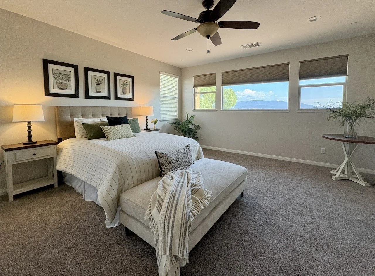 A minimalist bedroom with a large bed, beige upholstered headboard, striped bedding, and multiple pillows. On each side of the bed are matching nightstands with lamps. There is a window with brown blinds showing a view of mountains and blue sky. A ce