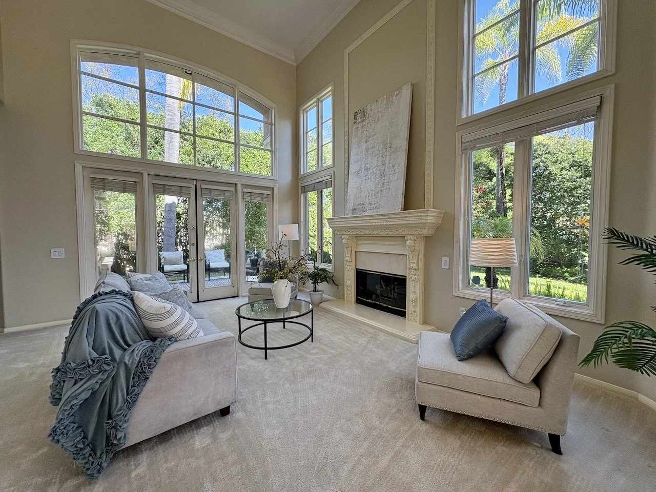 Bright living room with large arched and rectangular windows showing green trees outside, a white fireplace with decorative detailing, beige furniture, and a glass coffee table.