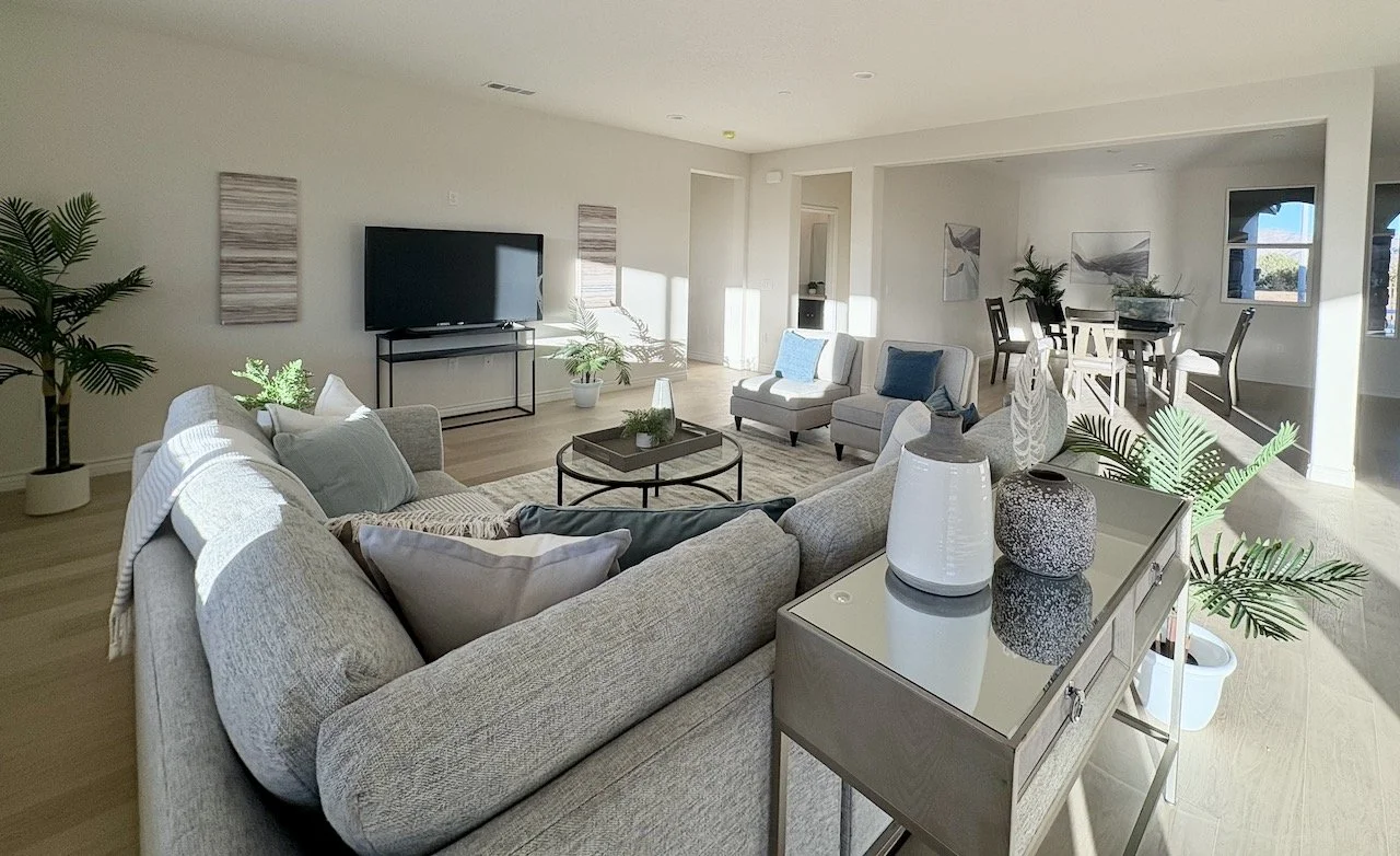 Bright living room with beige sofa, armchair, coffee table, and plant decor, adjacent to a dining area with chairs and tables, and a large flat-screen TV on a stand.