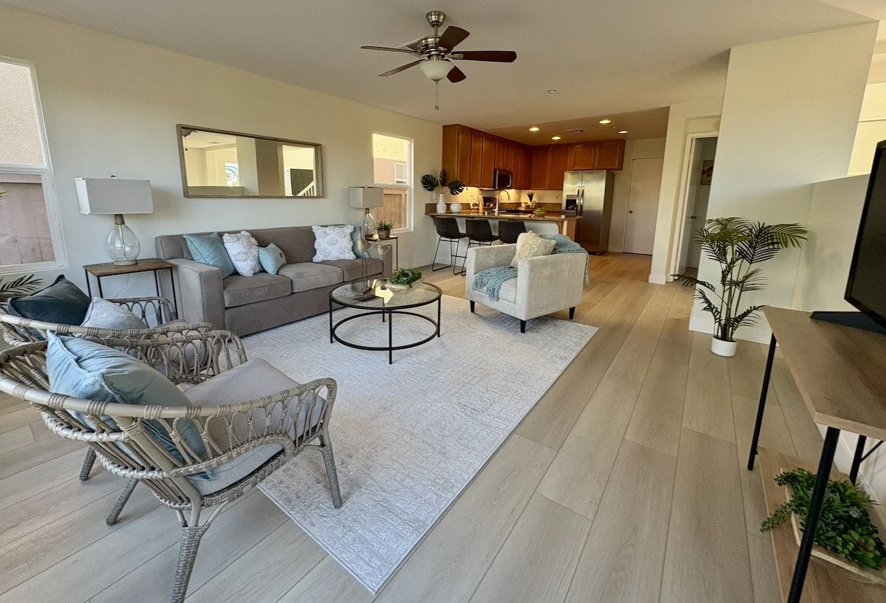 Open-concept living room with a gray sofa, armchair, wicker chairs, and a coffee table, overlooking a kitchen with wooden cabinets and bar stools.