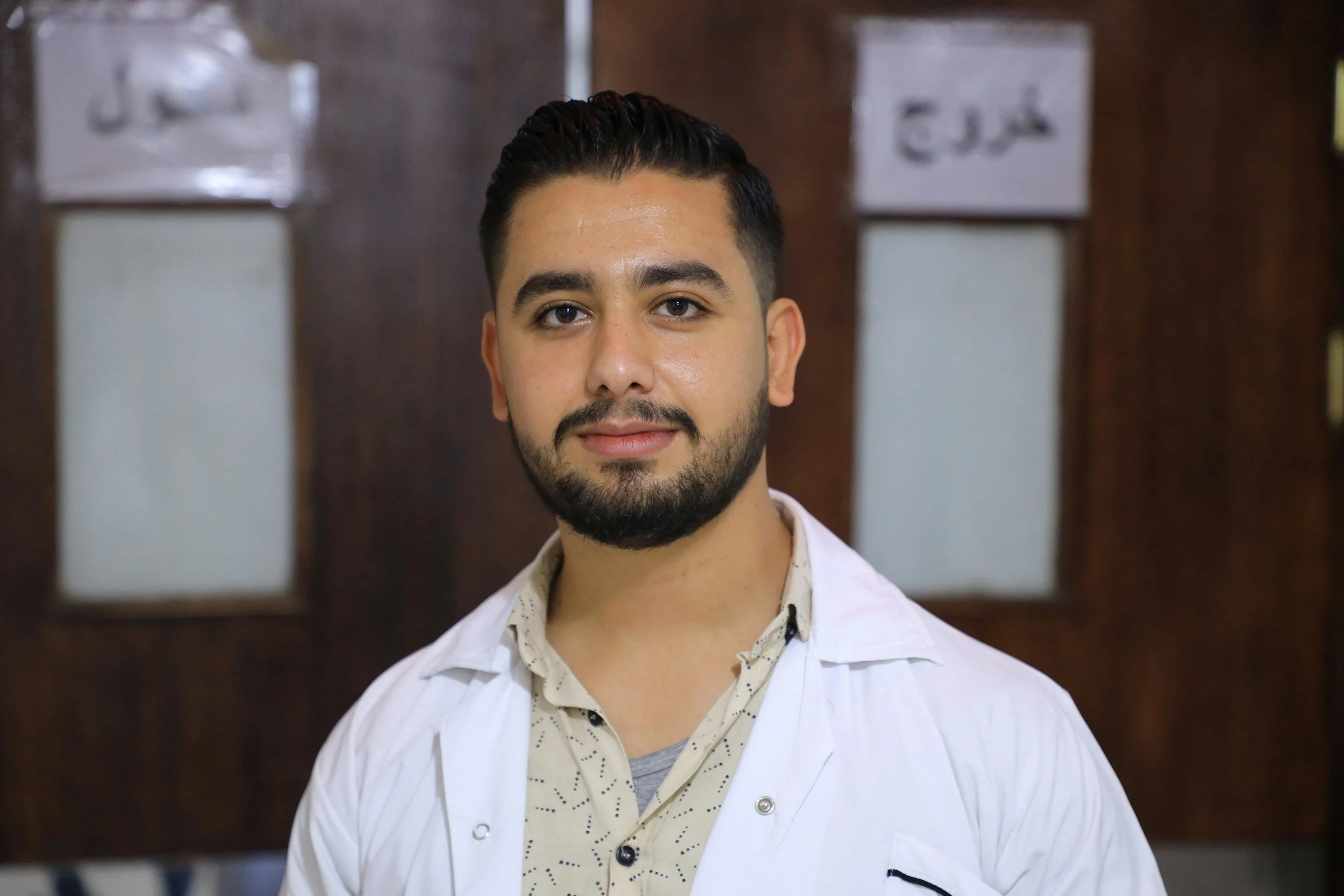A young man with dark hair, styled back, and a beard, wearing a white jacket over a patterned shirt, standing in front of a dark wooden background with framed pictures or documents.