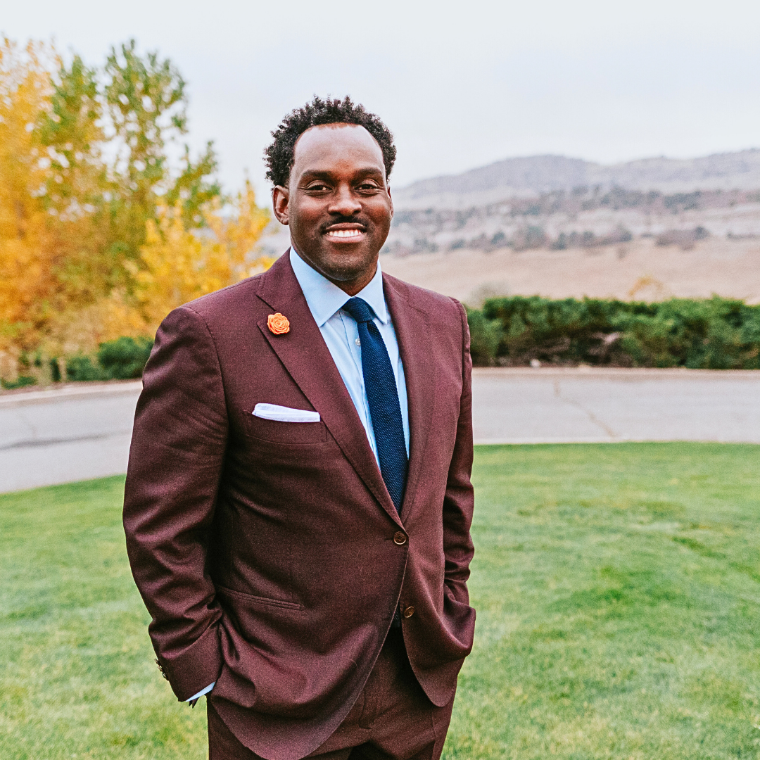 A man in a burgundy suit with a light blue shirt and dark blue tie standing outdoors on a green lawn, smiling with mountains and autumn trees in the background.