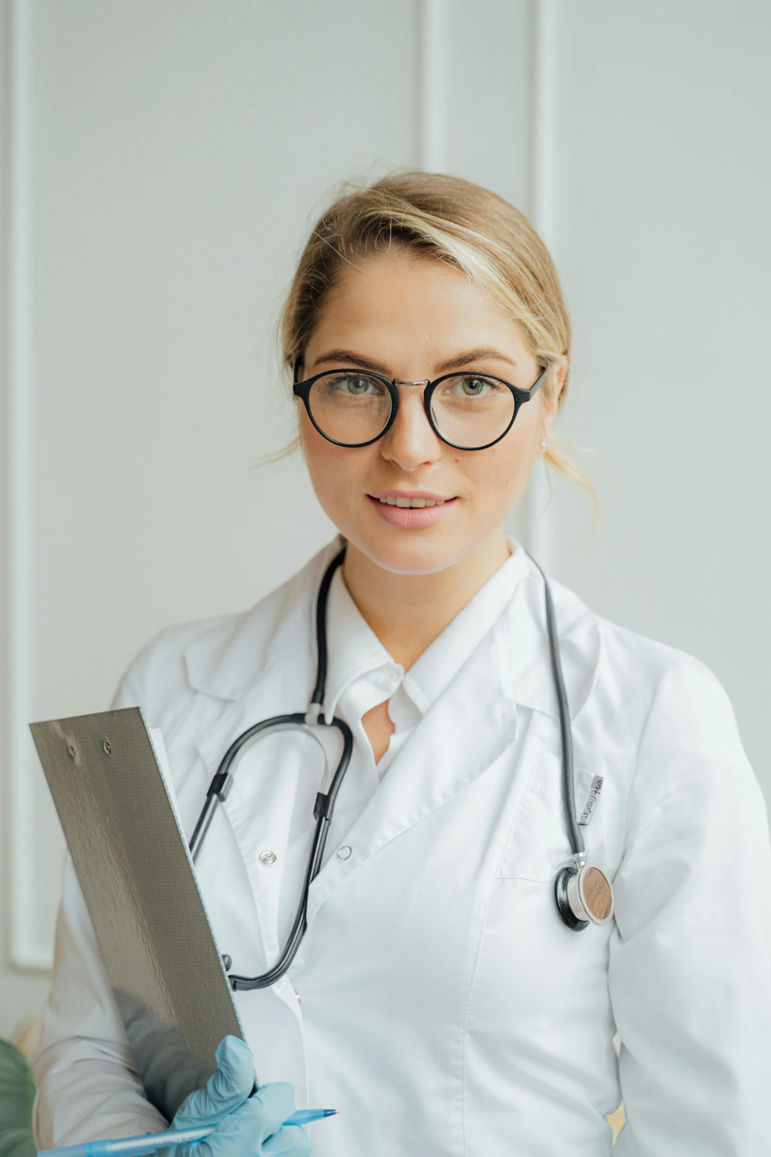 A female healthcare professional wearing glasses and a white lab coat, with a stethoscope around her neck, holding a clipboard and a pen, in a medical setting.