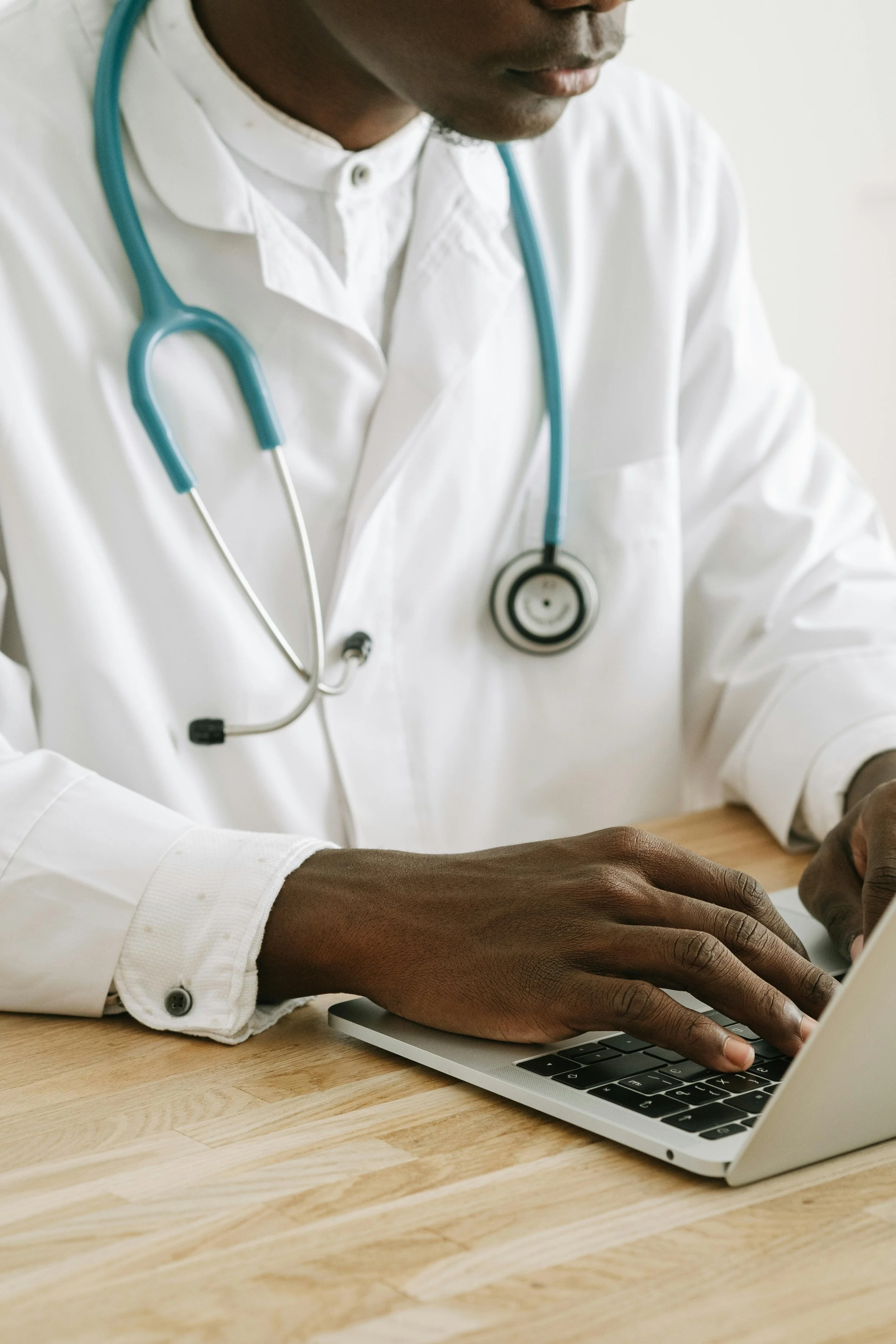 Close-up of a person with dark skin wearing a white shirt with buttoned cuffs and a stethoscope around their neck, working on a silver laptop at a wooden desk.