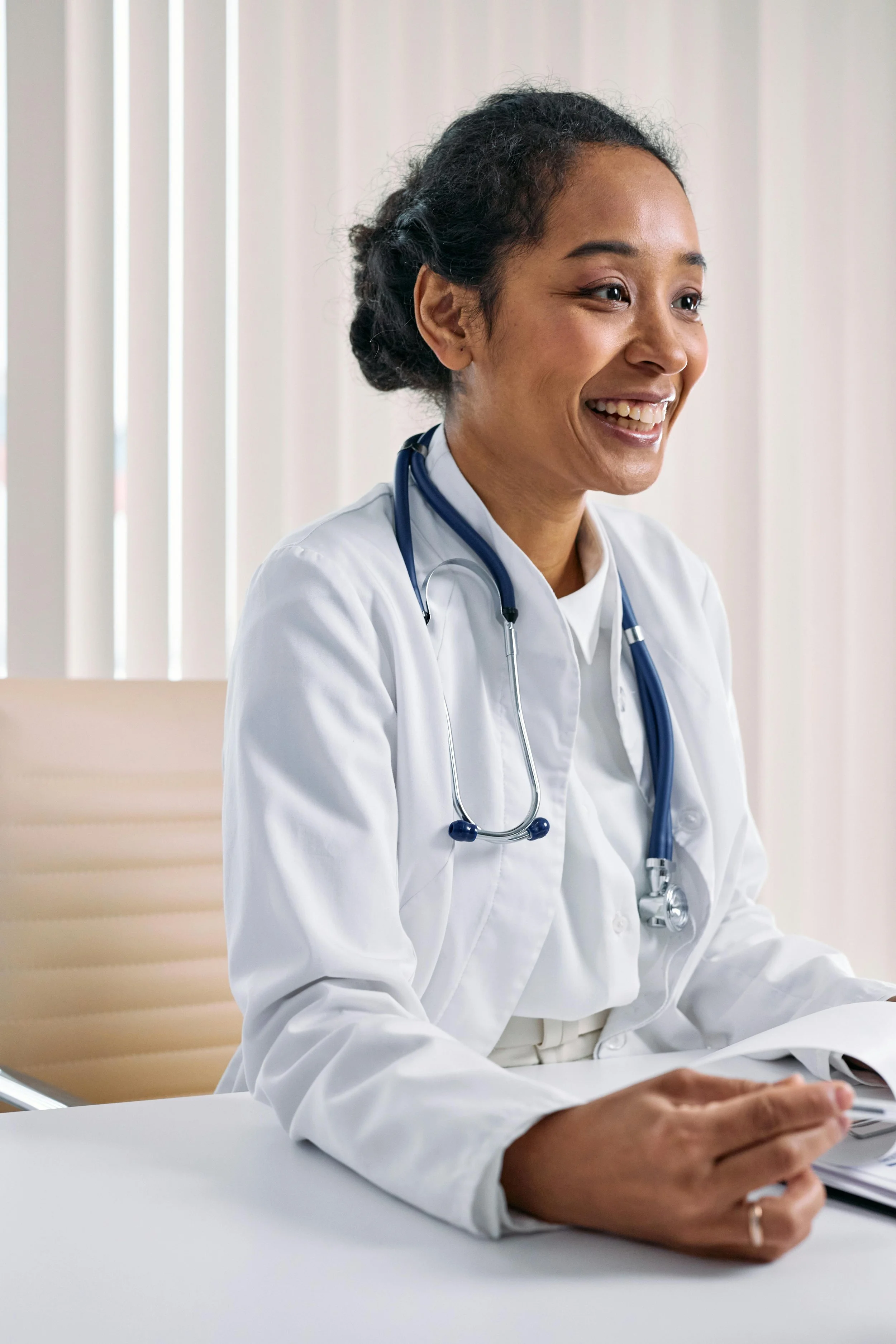 A female doctor smiling, wearing a white coat and stethoscope, sitting at a desk with hands resting on papers.