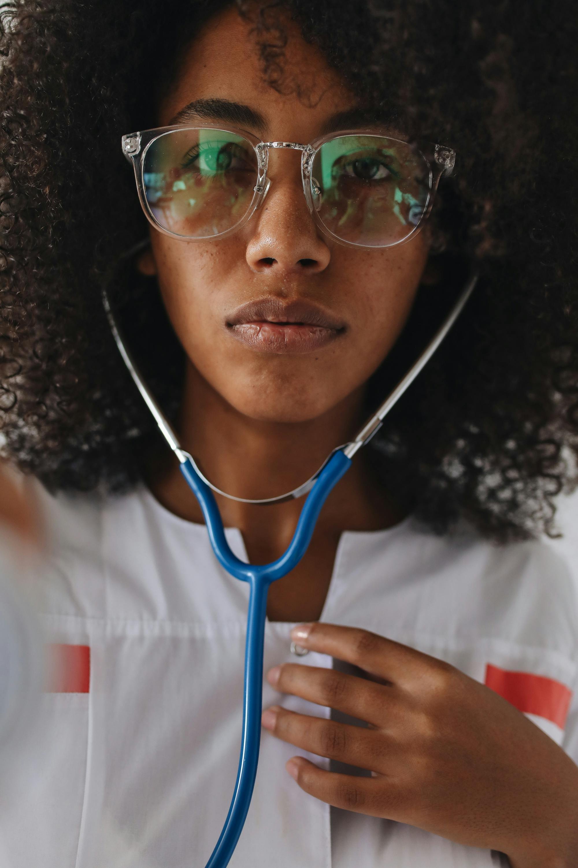 A woman with curly hair wearing clear glasses and a white shirt with red stripes, holding a blue stethoscope near her chest.