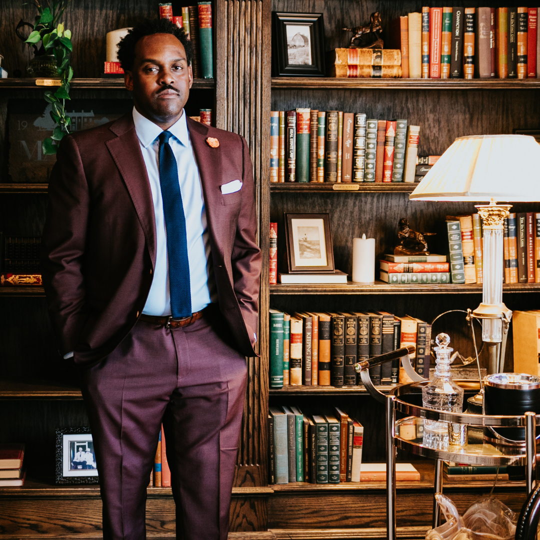 A man in a burgundy suit, light blue shirt, and navy tie standing with hands in pockets in a library filled with books and framed photos. A table with a lamp, glass decanter, and other items is nearby.