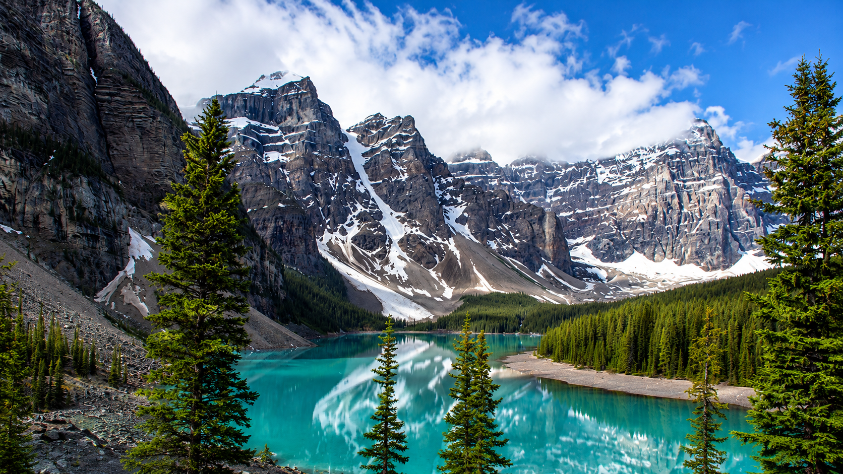 Snow-capped mountains reflecting in a turquoise lake surrounded by dense green pine trees.