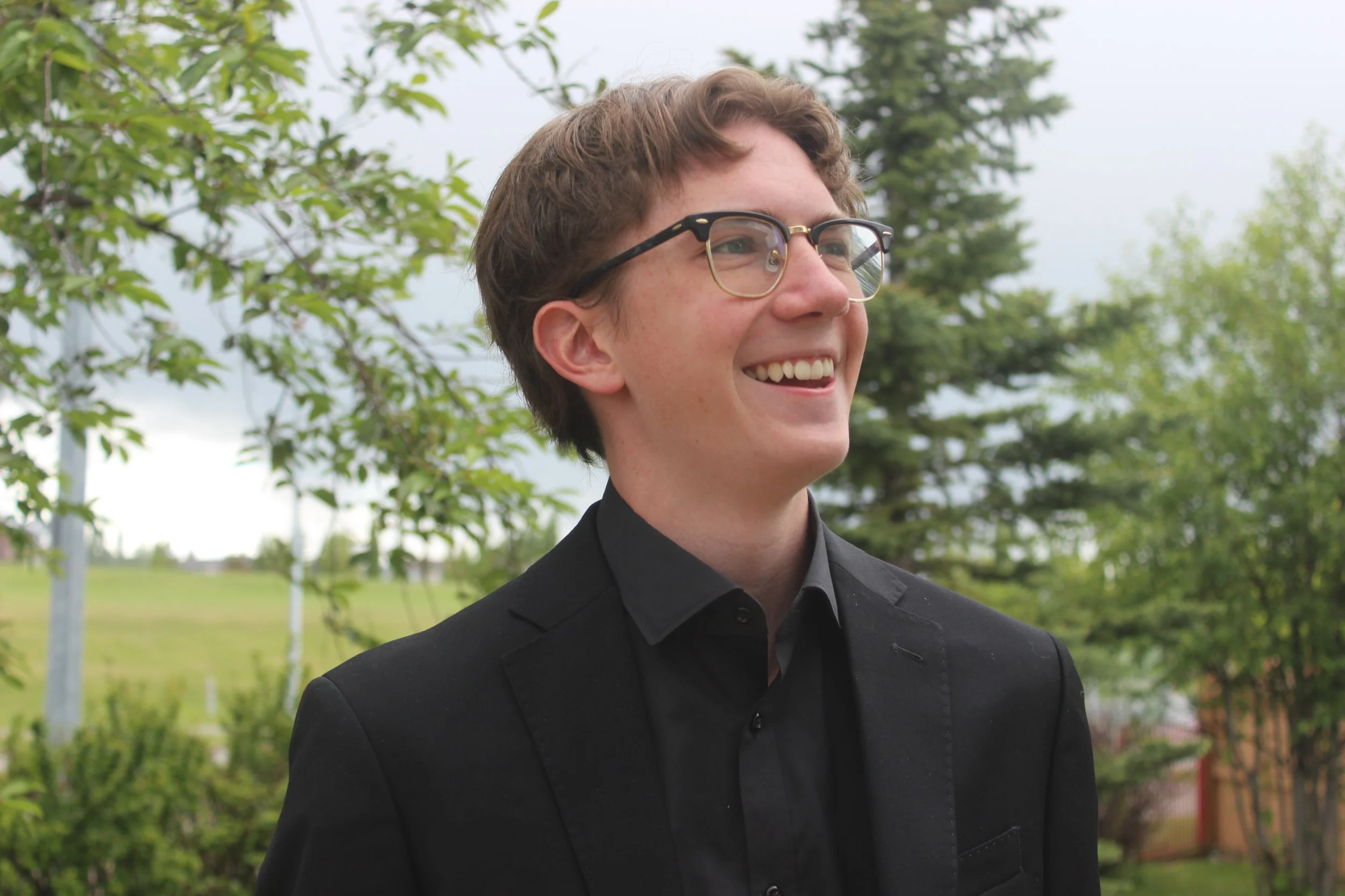 Young man with short brown hair, glasses, and a broad smile, wearing a black suit and black shirt, standing outdoors amidst green trees and a cloudy sky.