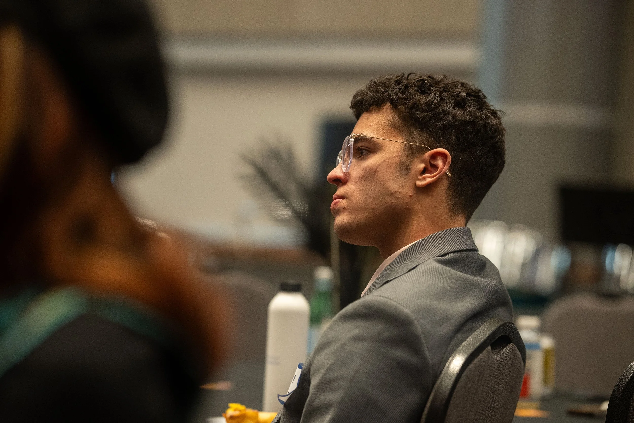 A young man with curly hair and glasses in a gray suit sitting in a conference room.