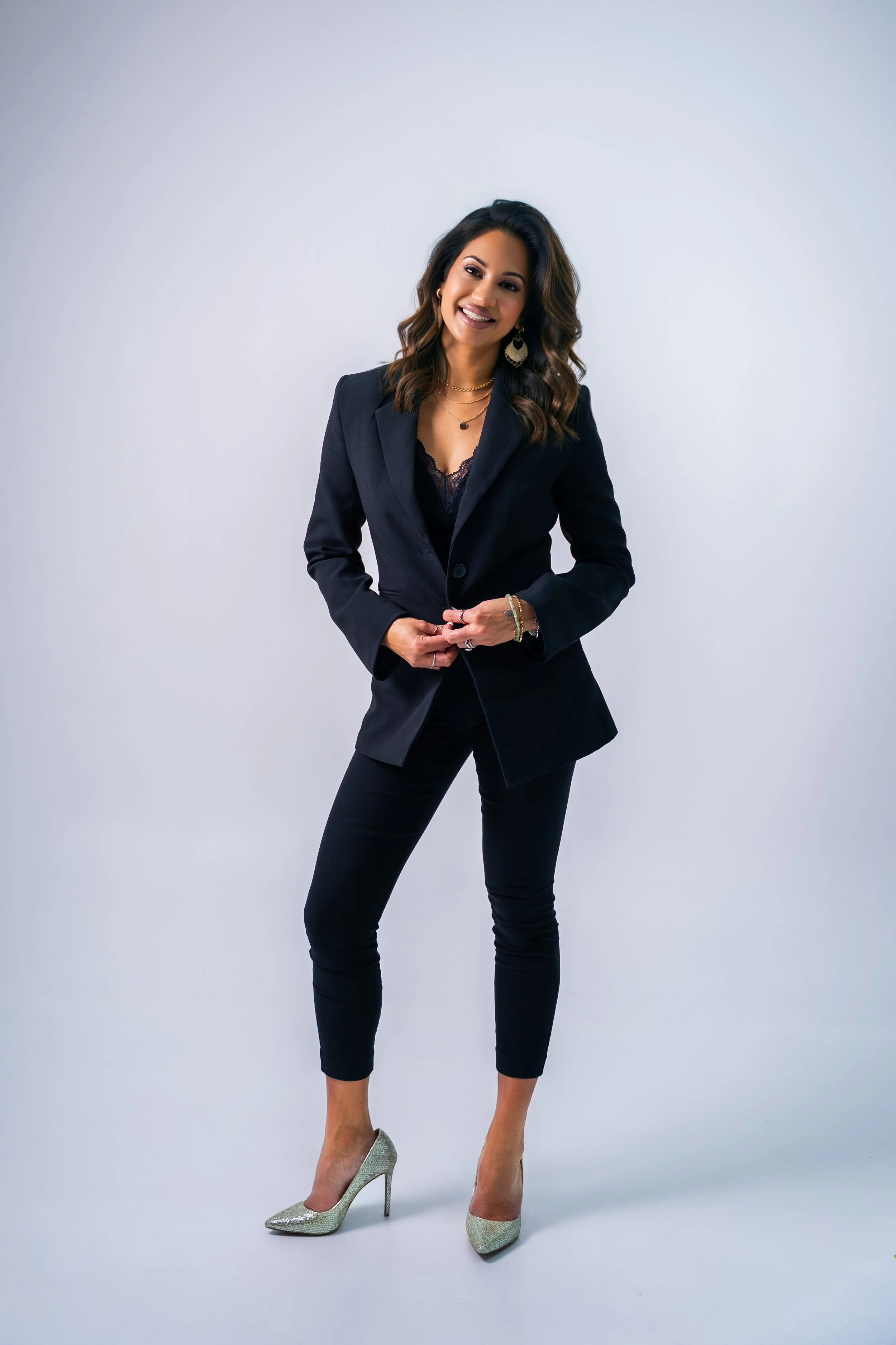 A woman dressed in a black blazer, black pants, and glittering high heels standing against a plain white background. She has shoulder-length wavy dark hair, earrings, and layered necklaces, and is smiling while posing with her hands clasped.