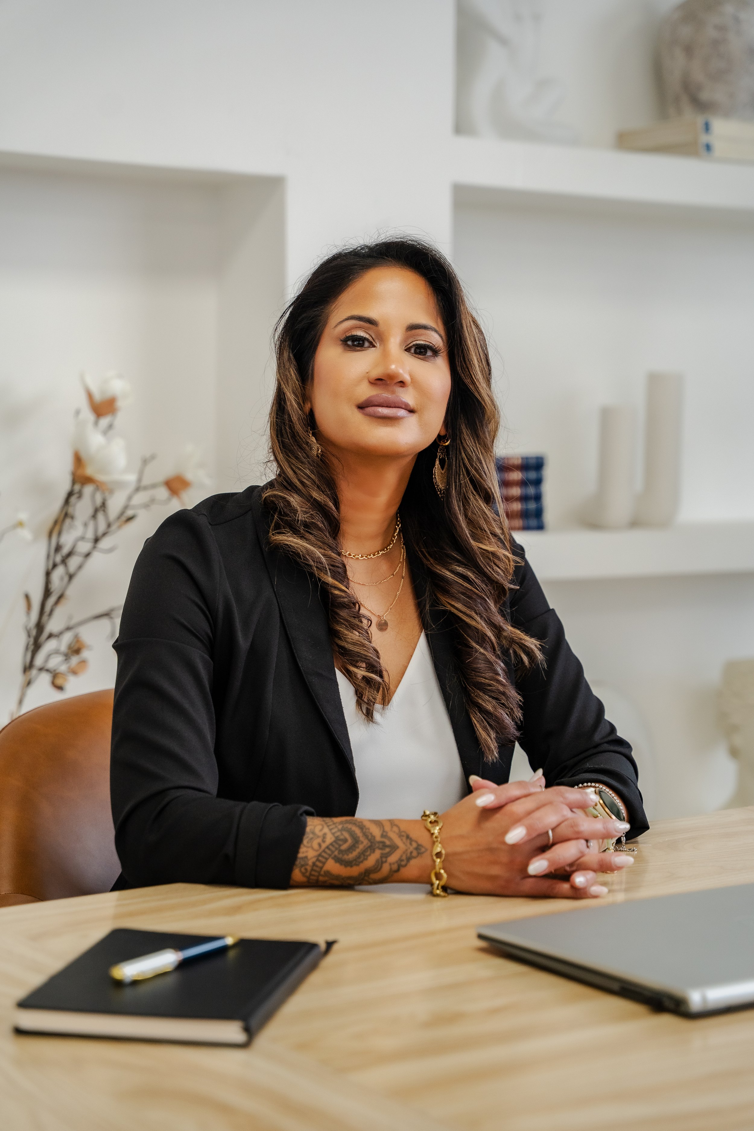 A woman with long, wavy hair and tattoos on her left forearm, dressed in a black blazer, sitting at a wooden table with notebooks, a pen, and a laptop, in a minimalist office with white walls and decorative shelves.