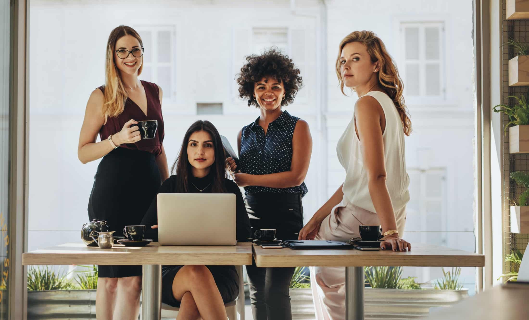 Five women gathered around a table in a bright, modern office space, with one woman seated at a laptop, and the others standing nearby, smiling and holding coffee mugs.