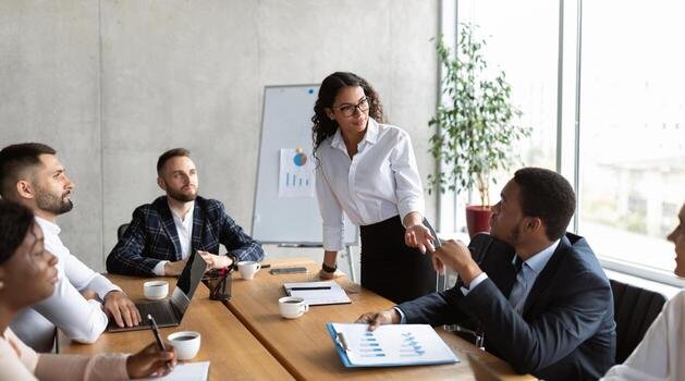 Woman standing and handing a business card to a man seated at a conference table in a modern office conference room with other colleagues.