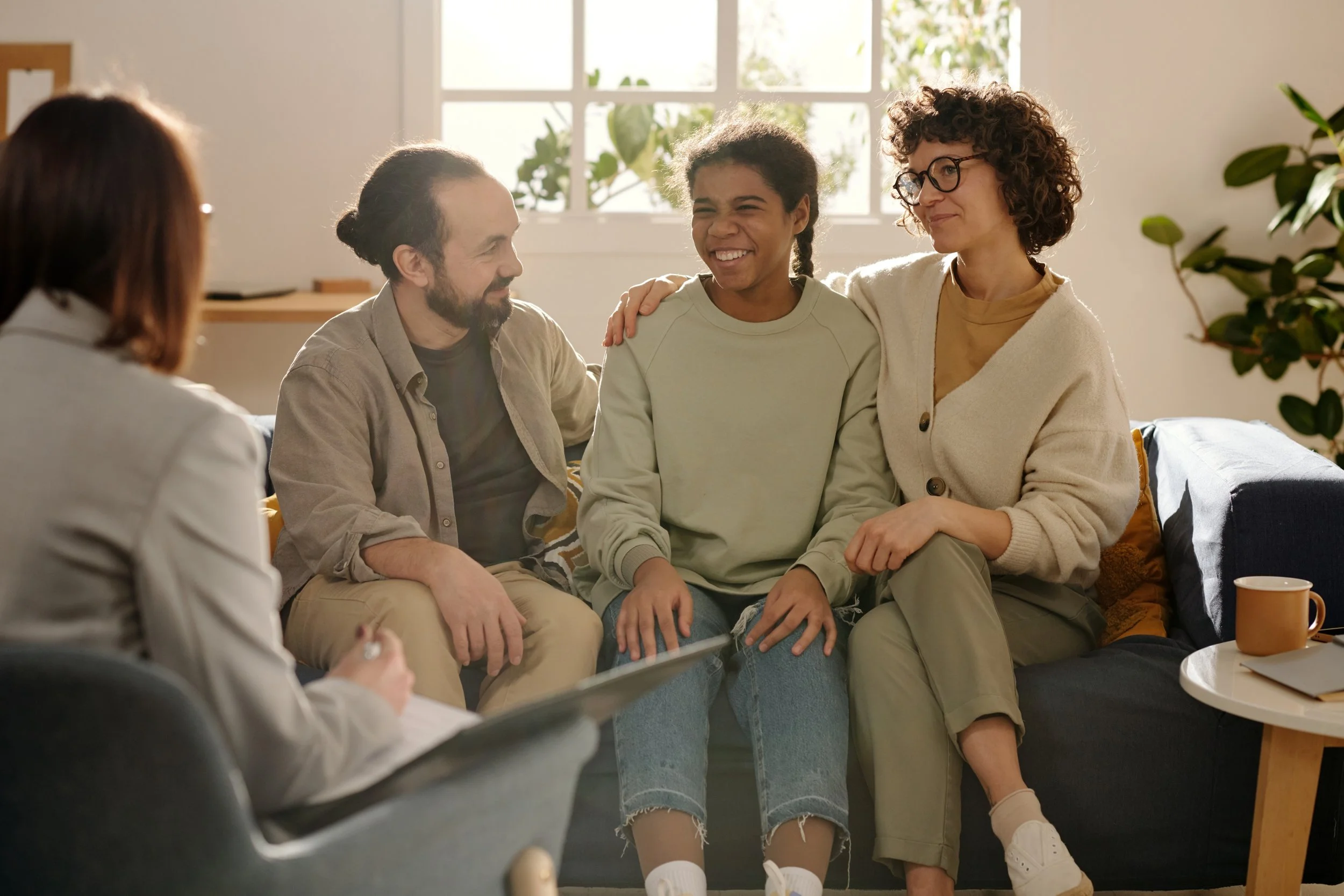 A group of five people sitting on a sofa in a well-lit room, engaged in conversation and smiling. The group includes a man with a beard, a young girl with braids, a woman with curly hair and glasses, and two other women. The setting appears cozy and warm, with plants and sunlight streaming through the windows.