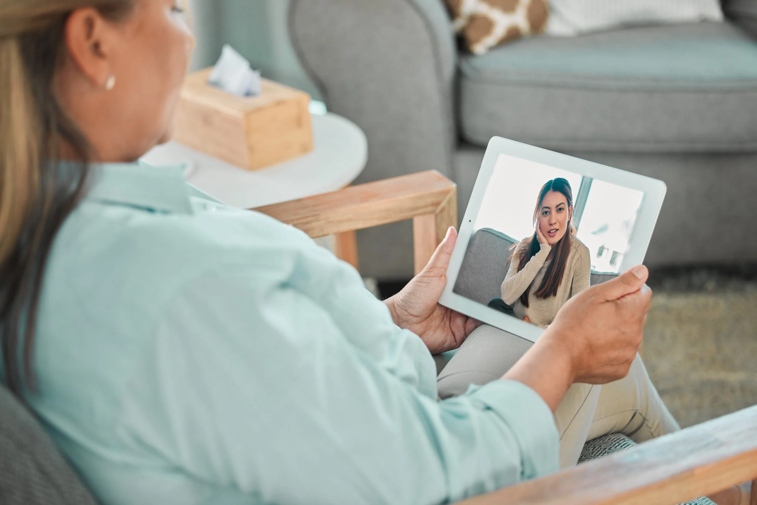 A woman holding a tablet with a video call of another woman touching her face.