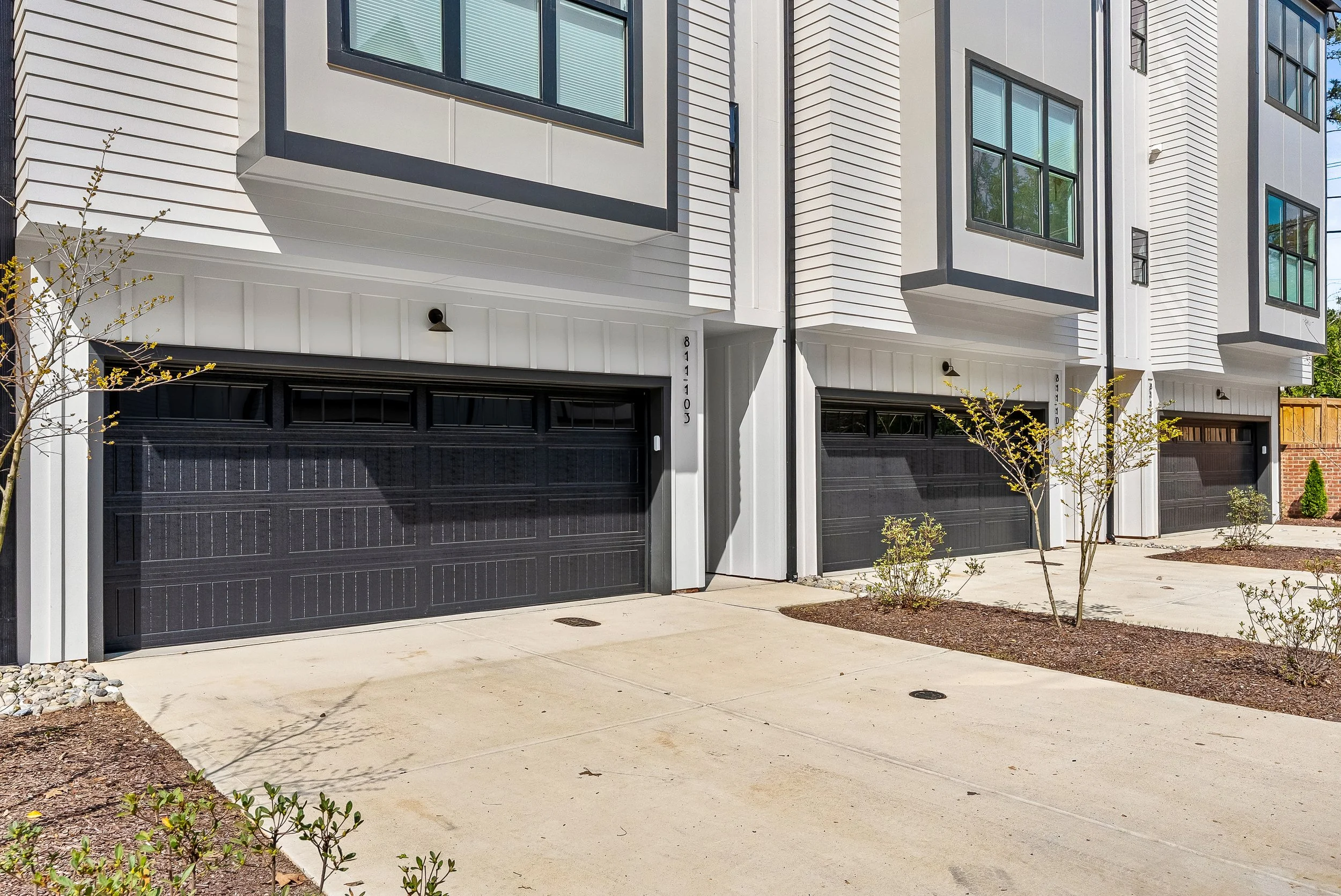 Front view of a modern townhouse complex with black garage doors, white siding, and large windows. Small trees and landscaping are visible in front.