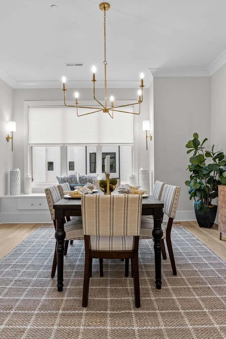 A modern dining room with a wooden table, eight patterned upholstered chairs, a gold chandelier, wall sconces, a large window with white blinds, a potted plant, and a rug.