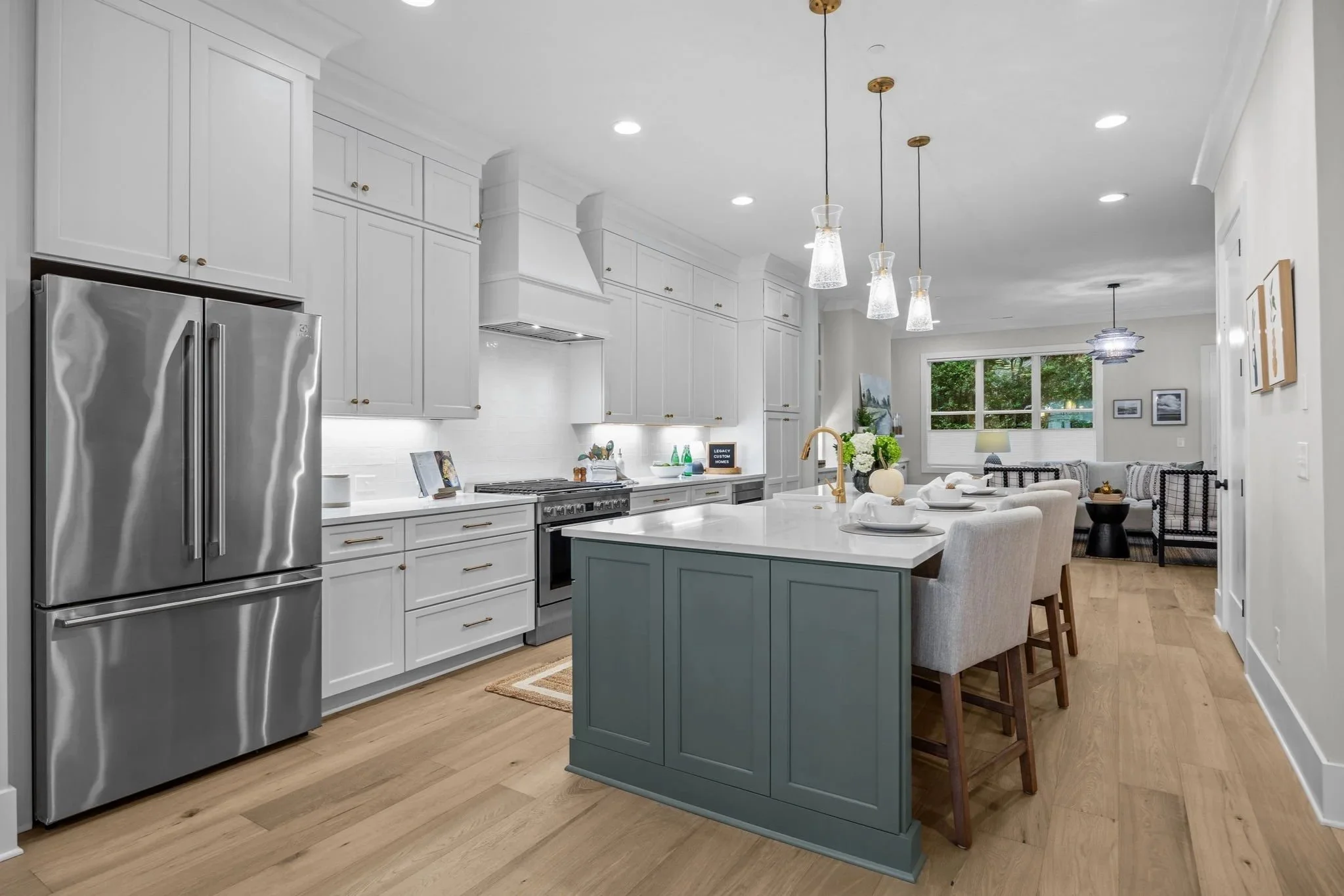 Modern kitchen with white cabinets, stainless steel refrigerator, island with a blue base, and hanging pendant lights. In the background, there is a living area with a window, artwork, and seating.