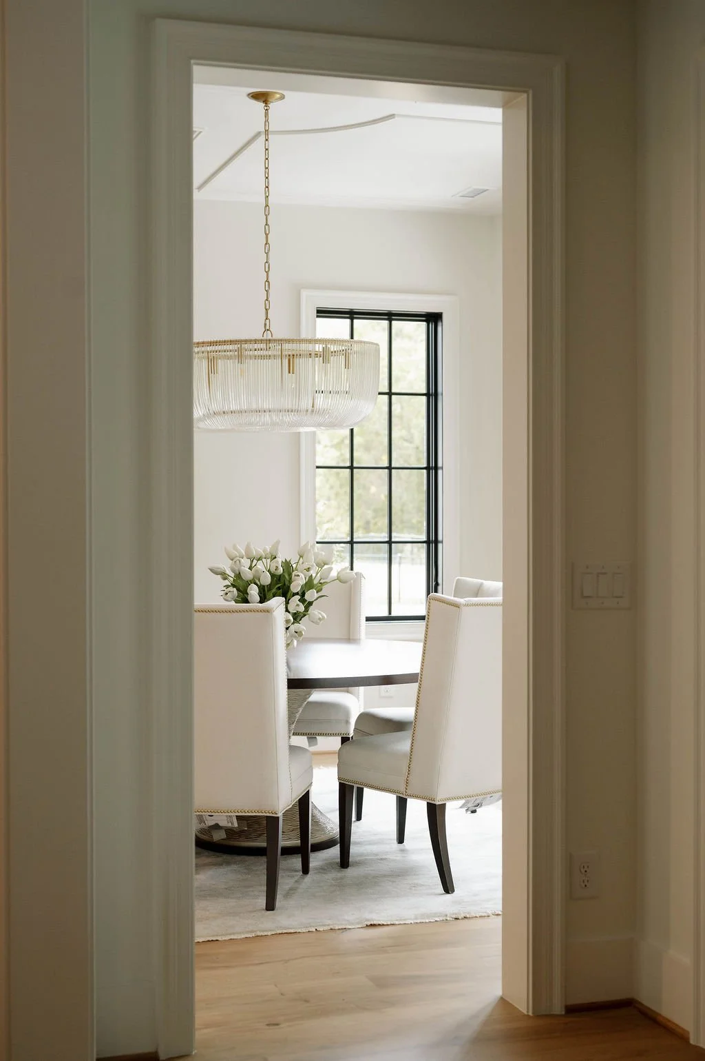 View into a bright dining room with a round table, white upholstered chairs, a flower arrangement, and a large window.