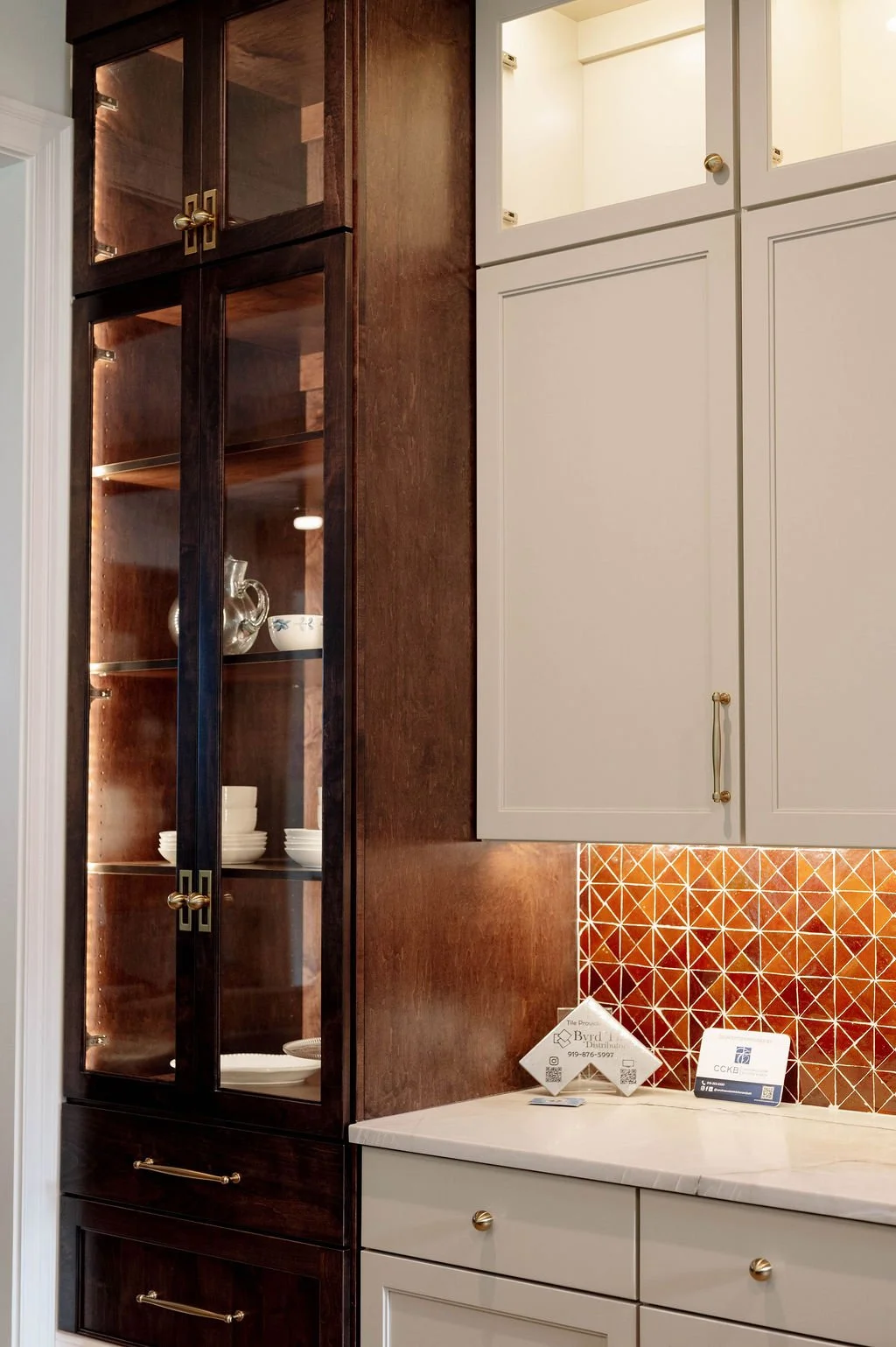 Kitchen with dark wood and white cabinets, glass display cabinet with dishes, copper hardware, and a geometric orange backsplash.