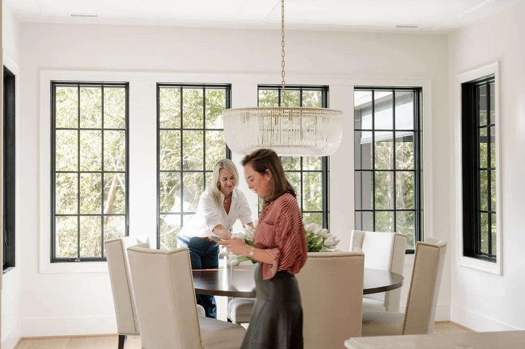 Two women in a bright dining room with large black-framed windows and a modern chandelier. One woman in a white shirt is adjusting a flower arrangement on a round dining table, while the other woman, in a reddish-brown blouse, is looking at her phone.