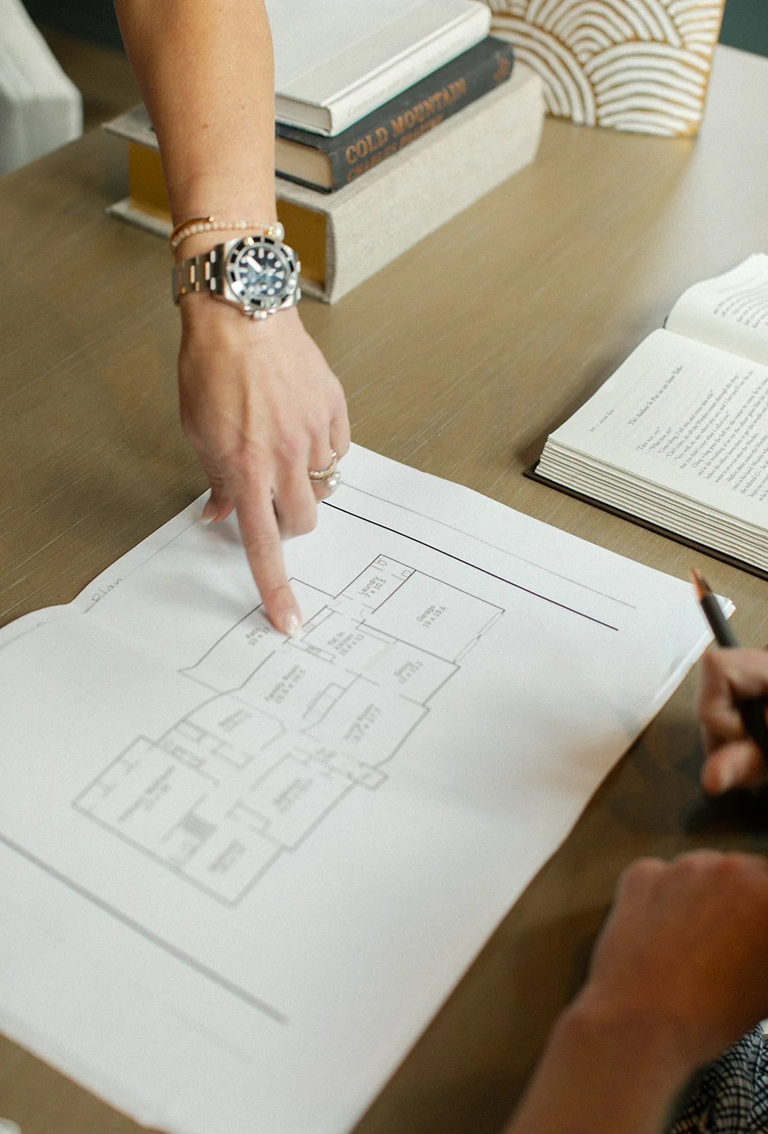 A person is pointing at a floor plan drawn on a large sheet of paper, likely discussing architecture or interior design, with a watch and bracelets on their wrist and an open book nearby.