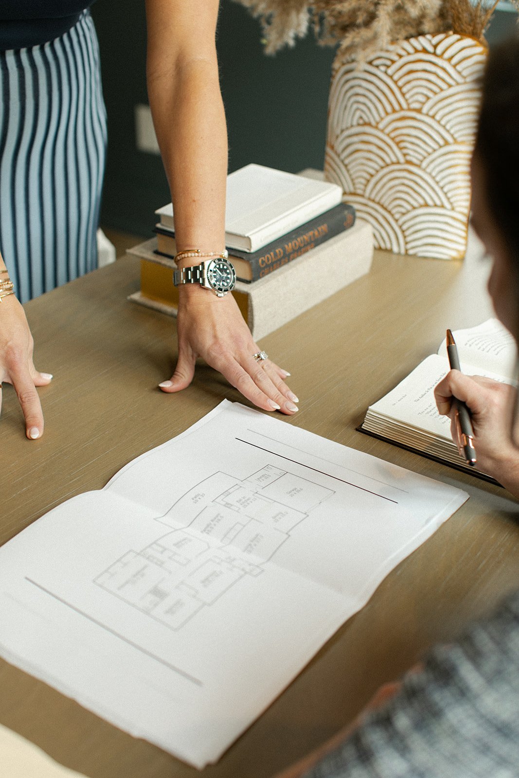 Two people are discussing a house plan at a wooden table, with blue-striped pants and watches visible. One person is pointing at the blueprint, and the other is holding a pen. Books and a decorative vase are on the table.