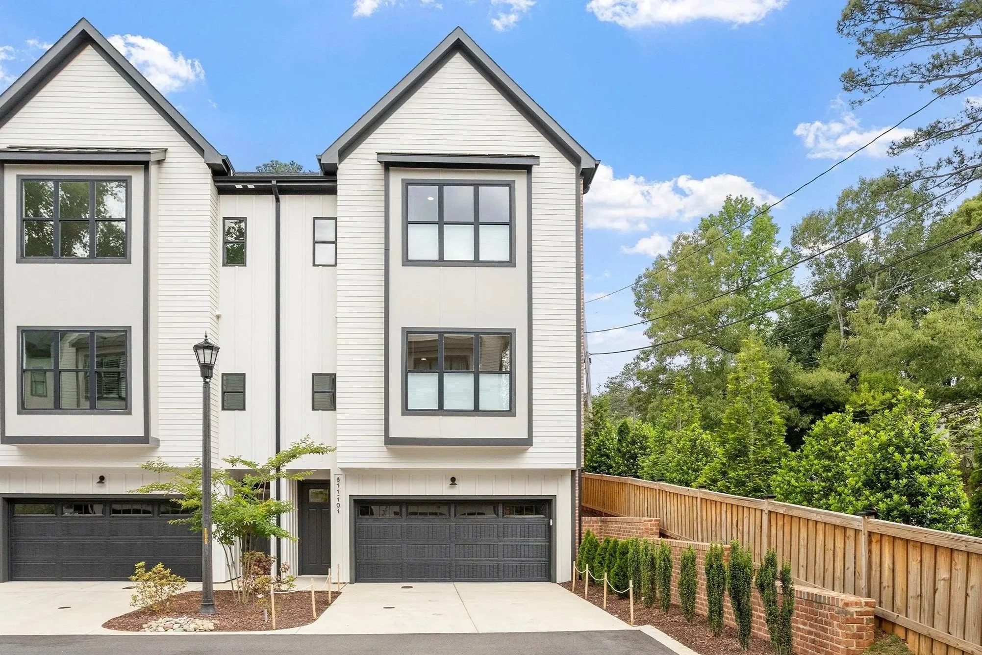 Front view of a modern multi-story house with black garage doors, white siding, and black window frames, surrounded by greenery and a wooden fence, under a partly cloudy sky.