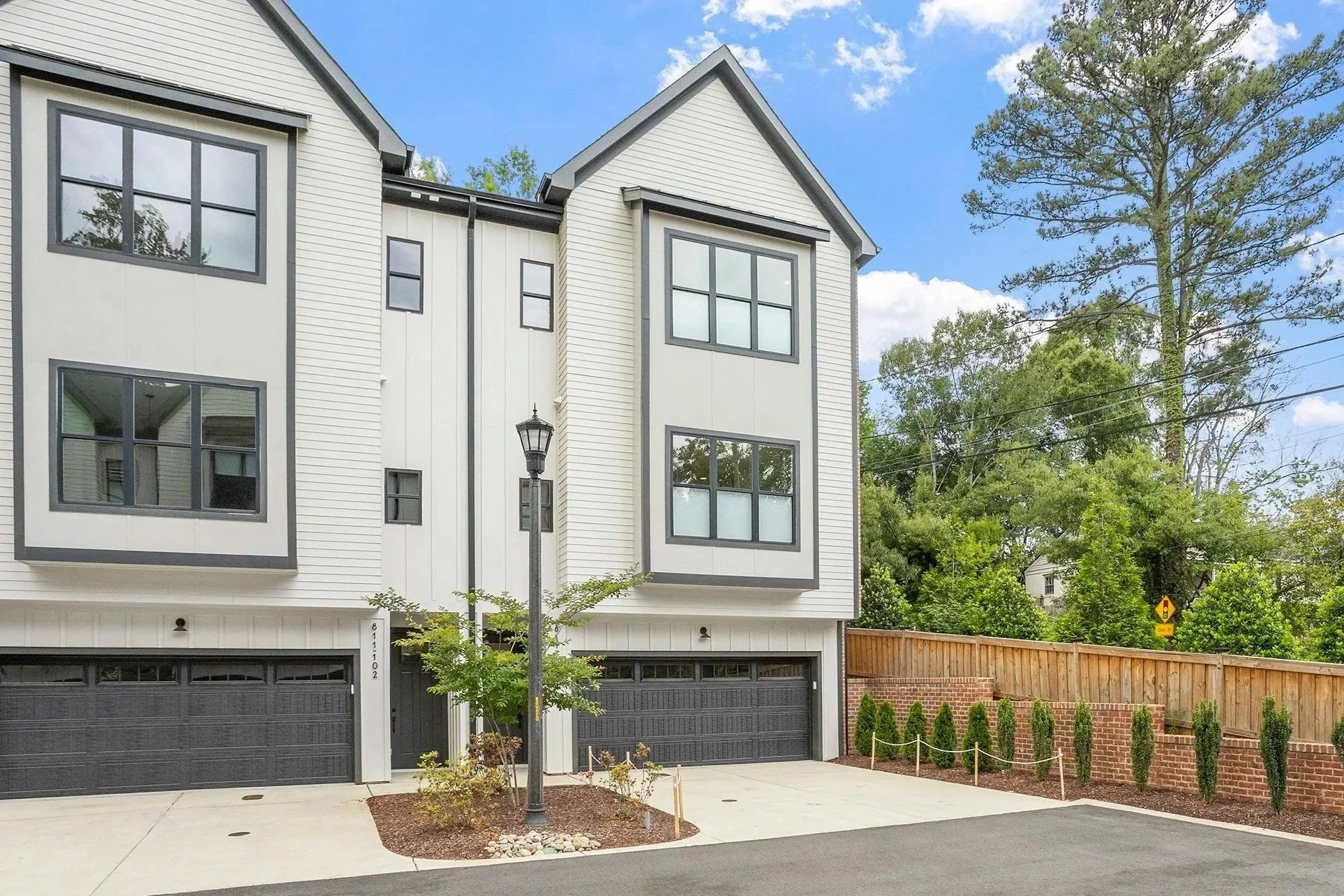 Modern townhouse with white siding, black window frames, and a black garage door, surrounded by green trees and a wooden fence, under a blue sky with some clouds.